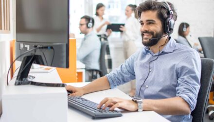 Customer support technician with headset working at computer in modern office. | i3solutions Support technician with headset providing IT assistance in a busy office environment.