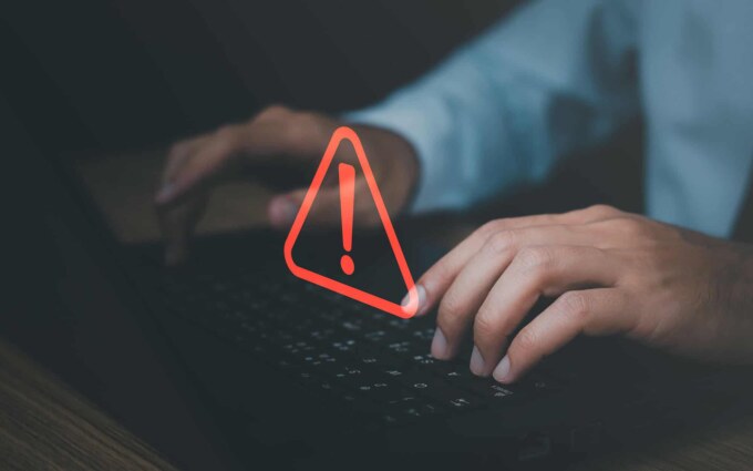 Side view of a mans hands hovering over laptop keyboard with red warning sign overlay