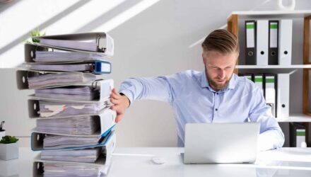 Data Management and Organization | i3solutions Man overwhelmed with stacked files and a laptop at a cluttered office desk.