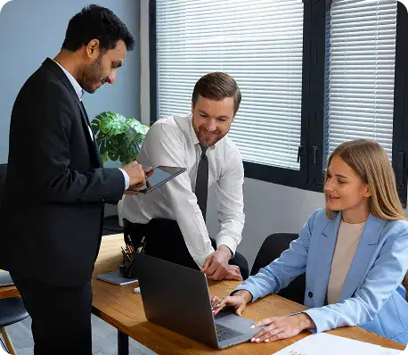 Professional Business Meeting with Laptops | i3solutions Three colleagues in business attire collaborate around a desk, one man showing a tablet while another types on a laptop as a third watches from the side.