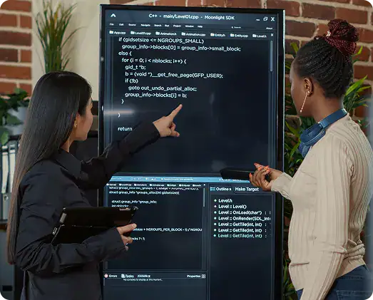 Software Development Collaboration | i3solutions Two women discuss code on a large screen displaying programming text in a brick-walled office setting.