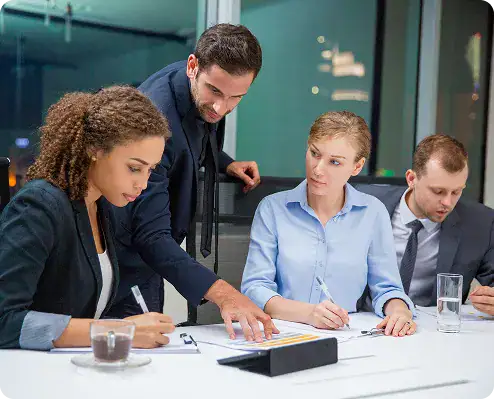 Professional Business Meeting for Data Solutions | i3solutions A man standing and pointing at documents while three colleagues sit at a white table, reviewing papers in a modern office conference room.