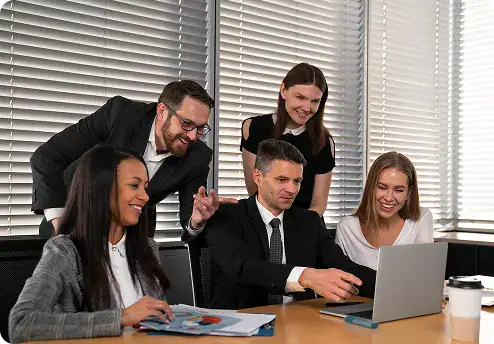 Team Collaboration for Dataverse Projects | i3solutions Four professionals collaborate around a laptop in a bright office, reviewing charts and documents on the table.