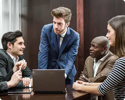 Team Meeting for Data Solutions | i3solutions Four professionals in business attire lean over a laptop, discussing a project in a conference room.