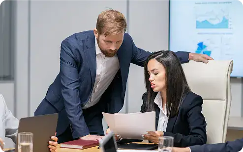 Business Meeting for Agile Teams | i3solutions Man in a blue suit leans over a woman in a white shirt, reviewing documents at a conference table.