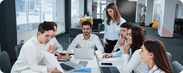 Team of Developers Collaborating in Office | i3solutions Team of six coworkers around a conference table with laptops and documents, collaborating in a bright open office.