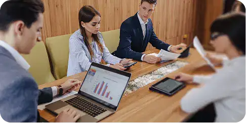dotnet-fixed-scope-initiatives A team of professionals around a conference table reviewing charts on a laptop and documents during a meeting.