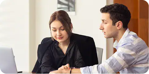 dotnet-hybrid-advisory-delivery-models Two professionals in an office discussing at a laptop, with the woman taking notes while the man points at a document or screen behind the laptop