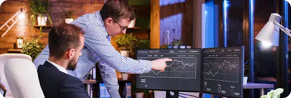 Data Analysis for .NET Application Development | i3solutions Two colleagues in a tech office review financial graphs on three monitor screens, one pointing at data on the central display.