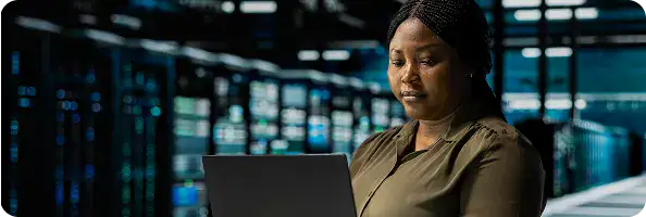 IT Professional Using Laptop in Data Center | i3solutions Professional woman using a laptop in a data center, with server racks and blue-lit equipment in the background.