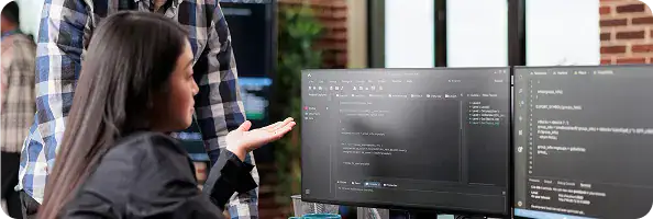 Software Developer Discussing Code | i3solutions Woman seated at a desk, discussing code with a standing teammate; dual monitors display dark code editors.