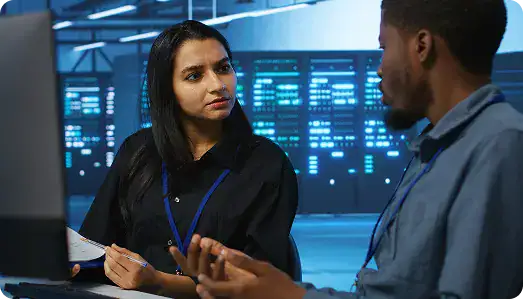 IT Professionals Discussing Data Security | i3solutions Two professionals discuss at a server room, woman with pen and blue lanyard listening to a man across a workstation