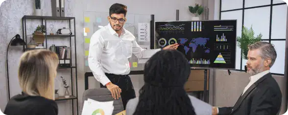 Team Collaboration in Agile Environment | i3solutions A presenter explains data to colleagues in a conference room, with charts and a world map on a large screen.