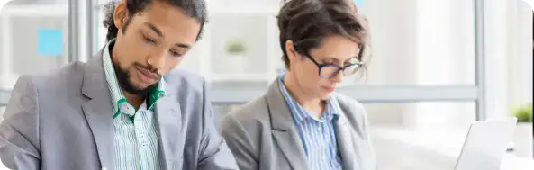 Business Professionals Collaborating in Office | i3solutions Two professionals in business attire work at a desk, looking down as they review documents or a laptop together in a bright office.