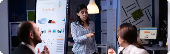 Team Collaboration in Portal Development | i3solutions Female presenter points at a tablet while two colleagues watch in a modern office with charts on screens and a whiteboard behind them.