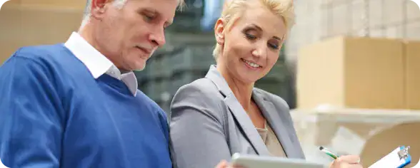 Team Collaboration in Agile Environment | i3solutions Two coworkers review a clipboard in a warehouse with boxes in the background.