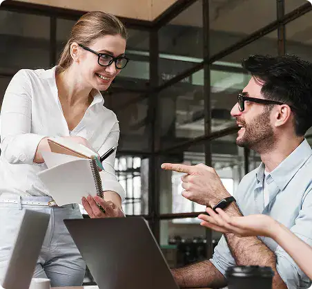 Professional Business Meeting with Two Colleagues | i3solutions Two colleagues collaborating in a modern office: a smiling woman in white with a notebook, and a man in glasses pointing while working on a laptop step-by-step discussion.