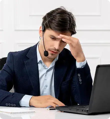 Frustrated IT Support Specialist | i3solutions Young man in a navy suit with a headset, resting his forehead while working on a laptop.
