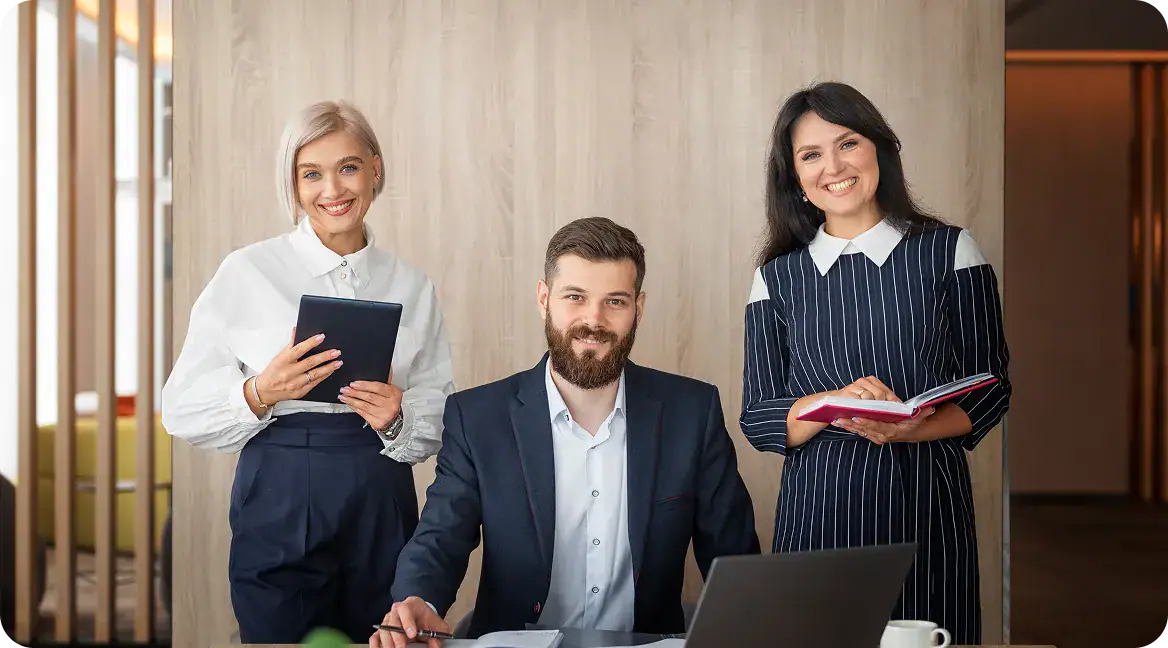 hybrid-transformation-knowledge-transfer-models | i3solutions Three business colleagues pose in a modern office: a man sits at a desk with a laptop, flanked by two women—one holding a tablet and the other a notebook—smiling at the camera.