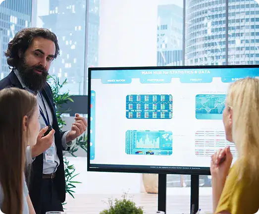 Business Data Analytics Presentation | i3solutions Bearded man presenting data dashboards on a large monitor to two colleagues in a bright office, gesturing toward the screen.