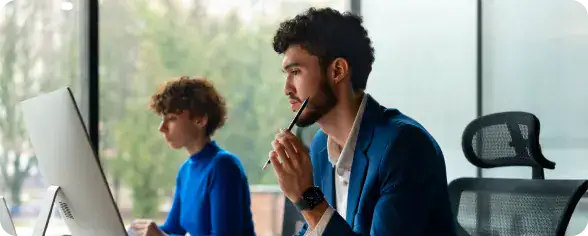 long-term-embedded-analytics-teams. | i3solutions Office scene with two workers at desks and desktop computers; foreground man in a blue blazer holds a pen to his lips, thinking.