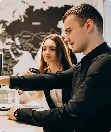 Business Meeting with Tech Focus | i3solutions Two coworkers at a desk, man in foreground smiling and reaching forward while a woman assists in the background, with a world map mural on the wall behind.