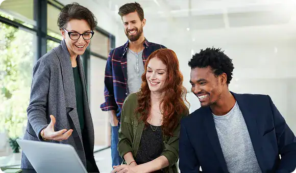 Team Collaboration in Modern Office | i3solutions Four diverse colleagues gathered around a laptop in a bright office, smiling and discussing the screen; woman with glasses gestures toward it.