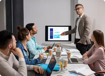 Professional Business Meeting Presentation | i3solutions Presenter in a blazer points to a chart on a monitor as colleagues listen around a conference table with laptops and notebooks nearby.