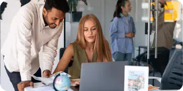 Team Collaboration in Portal Development | i3solutions Two coworkers collaborating in an office: a man leans over a desk toward a woman using a laptop, with papers and a globe on the desk.
