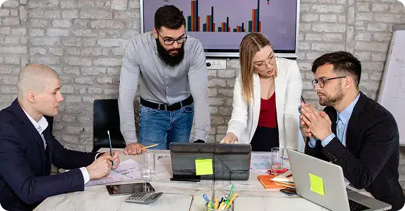 Dedicated Agile Team Collaboration | i3solutions Four professionals collaborate around a table, looking at a tablet and monitor displaying charts in a modern office.