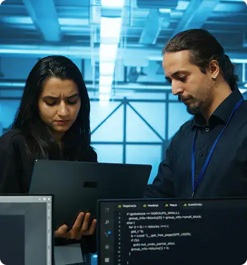 IT professionals collaborating on device | i3solutions Two IT professionals review code on a laptop in a data center with blue lighting.
