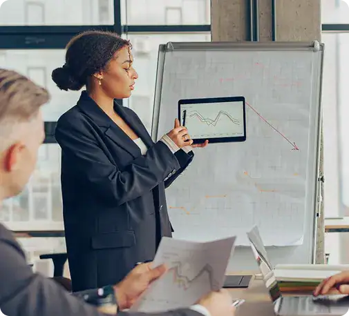 Businesswoman Presenting Data Analysis | i3solutions Professional woman presenting financial charts on a tablet to colleagues in a boardroom with a whiteboard displaying graphs in the background.