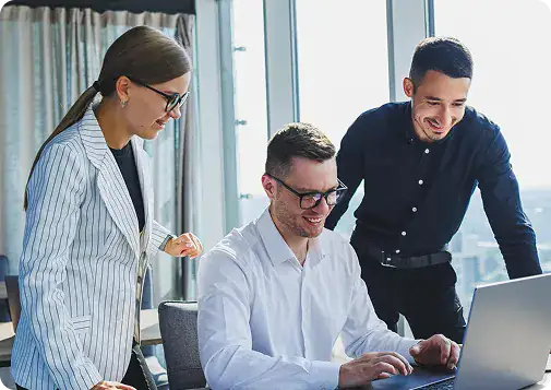 Team Collaboration for IT Solutions | i3solutions Three coworkers collaborate around a laptop in a bright office space with one standing woman, one seated man, and another man leaning in.
