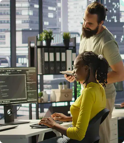 IT Collaboration in Modern Office | i3solutions Bearded man standing beside a seated woman at a computer, reviewing code on a monitor in a bright office.