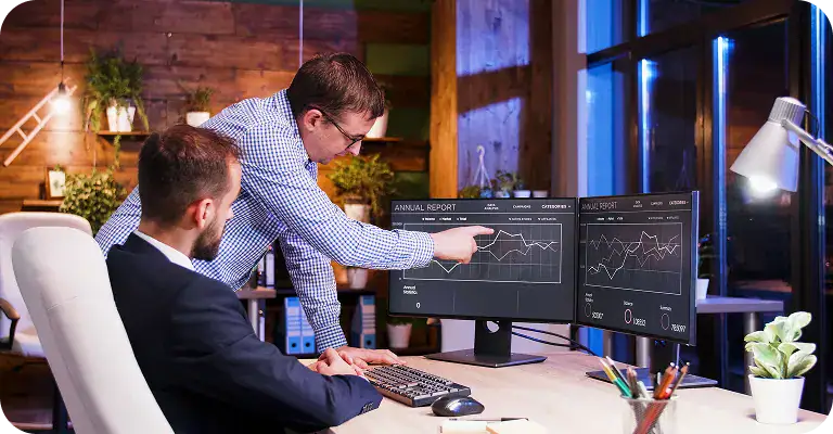 Business Analysts Reviewing Data Analytics | i3solutions Two professionals analyzing data charts on dual monitors in a modern office.