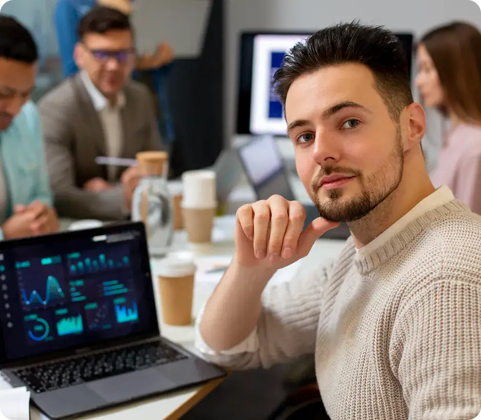 Young man in a business meeting with colleagues and data charts on laptop and screens.