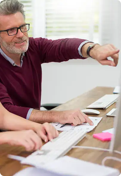 i3solutions Business Meeting Bearded man with glasses in a burgundy sweater points toward the computer screen as colleagues type at a shared desk.