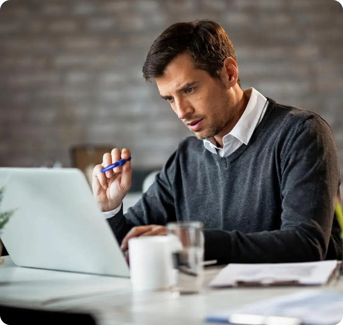 Focused man working on laptop with pen in hand in a modern office.