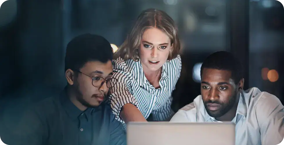 Team Collaborating on Data Analytics | i3solutions Three colleagues huddle around a laptop, focused on the screen during a night shift in a modern office environment, collaborating intently.