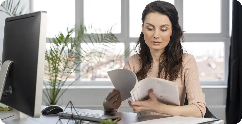 Businesswoman Analyzing Data Reports | i3solutions Woman seated at a desk, reviewing documents with a computer monitor nearby and a plant in the background.