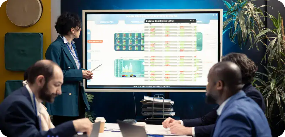 Business Meeting with Data Presentation Woman in a teal blazer presents beside a large screen filled with charts while four colleagues sit around a table with laptops and coffee cups, plants in the background.