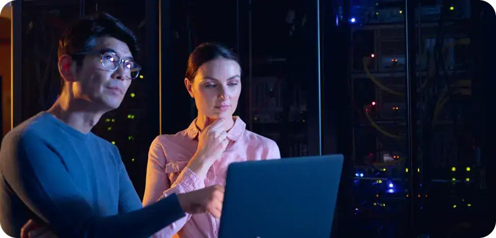 IT professionals working on a laptop Two IT professionals stand beside a laptop in a data center, reviewing information on the screen amid server racks and glowing cables.