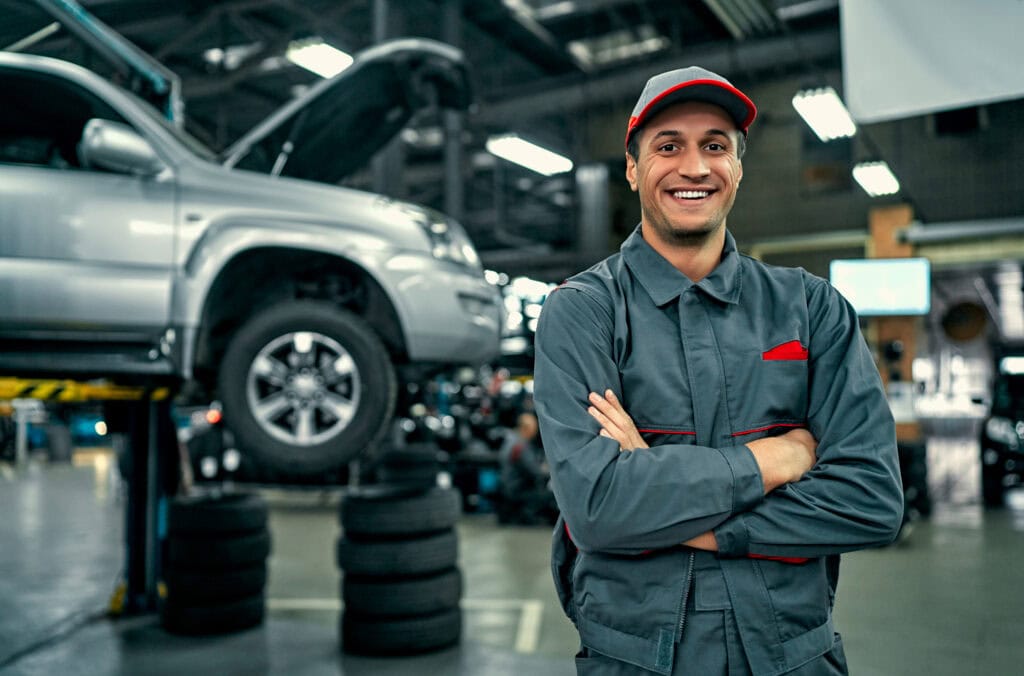 Automotive technician smiling in a car repair shop with a vehicle on a lift.