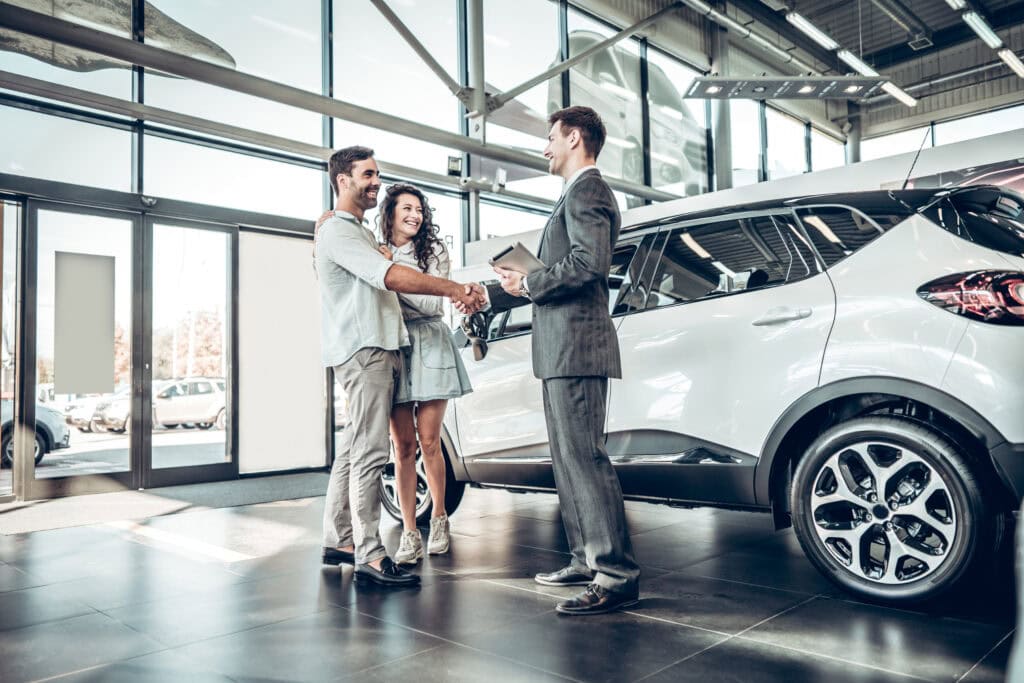 Happy couple shaking hands with car dealer in showroom with new vehicle.