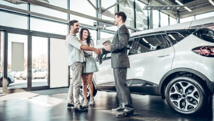 Happy couple shaking hands with car dealer in showroom with new vehicle.